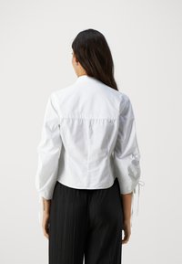 Camicia bianca con colletto, maniche a palloncino, tessuto strutturato, dettaglio arricciato sul retro e dettagli con nastri; indossata con pantaloni neri plissettati.