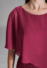 Blouse en mousseline bordeaux avec un large décolleté et des manches courtes flottantes. Associée à une jupe en dentelle texturée de couleur bordeaux foncé.