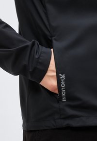 Χέρι που γλιστρά στην τσέπη με φερμουάρ ενός μαύρου μπουφάν Houdini, εμφανίζοντας τη μανσέτα του μανικιού και το λογότυπο Houdini κοντά στο φερμουάρ της τσέπης.