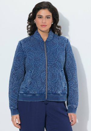 Frau trägt eine blaue, gesteppte Jacke mit Reißverschluss und abstrakten Mustern sowie passende blaue Hosen, steht vor weißem Hintergrund.