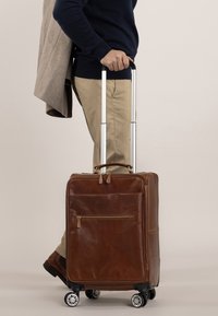 Brun l æderkuffert med en teleskopisk håndtag, fire hjul, en frontlomme med lynlås og et slankt design, båret mens man holder en frakke.