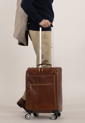 Maleta de cuero marrón con un mango retráctil, cuatro ruedas, un bolsillo frontal con cremallera y un diseño elegante, llevada mientras se sostiene un abrigo.