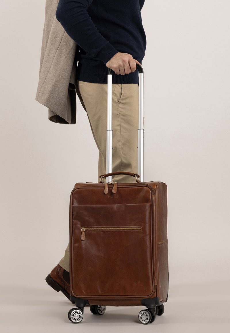 Brun læderkuffert med en teleskopisk håndtag, fire hjul, en frontlomme med lynlås og et slankt design, båret mens man holder en frakke.