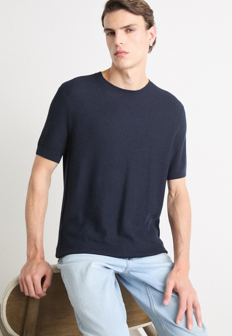 T-shirt a maniche corte blu navy in maglia con scollo rotondo, realizzato in tessuto strutturato. Indossato con jeans blu chiaro, appoggiato a una sedia di legno.