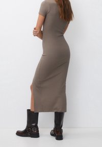 Robe midi beige à manches courtes, près du corps, avec une fente sur le côté, accompagnée de bottes en cuir noir brillantes montantes avec une boucle.