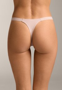 Ženska v bež tangicah, prikazana od zadaj in spodnjega dela trupa na enostavni podlagi.