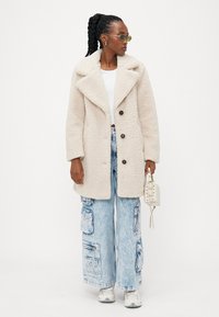 Beige shearling-takk med store rever og tre knapper, kombinert med lyseblå baggy jeans med lommer. Modellen holder en liten hvit håndveske.