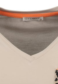 Top beige de escote en V con ribete naranja en el cuello, que presenta un ligero brillo. Incluye un bolsillo con un diseño de estampado de leopardo. Etiqueta de la marca visible.