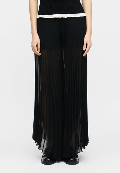 Γυναίκα που φοράει μαύρο λεπτό, πλισέ παντελόνι με φαρδιά πόδια πάνω από μαύρο σορτς, σε συνδυασμό με μαύρα παπούτσια και μαύρο τοπ με λευκή δαντέλα στο τελείωμα.