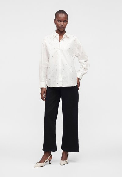 Femme portant un chemisier blanc à manches longues avec de petits motifs, un pantalon noir à jambes larges et des chaussures blanches à talons bas, debout sur un fond uni.