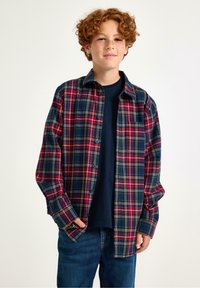 Rudy chłopiec w czerwonej, zielonej i żółtej koszuli w kratę, zapiętej na guziki, na granatowym t-shircie, w niebieskich dżinsach, stoi z rękami w kieszeniach.
