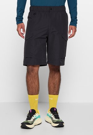 Homme portant un short cargo noir, des chaussettes jaunes Nike et des chaussures de course multicolores sur fond blanc.