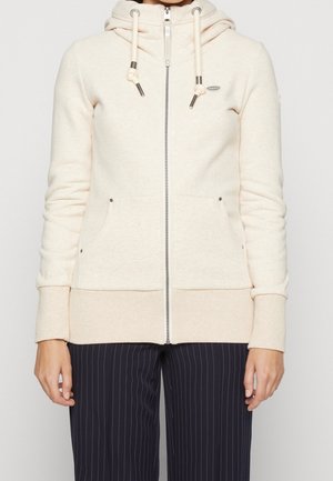 Person wearing a cream zip-up hoodie with drawstrings and side pockets, paired with black pinstriped pants.