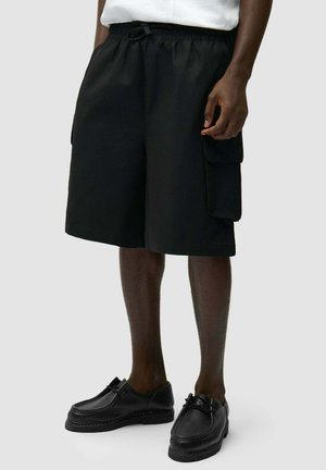 Pantaloncini cargo neri con tasche laterali indossati con scarpe nere stringate su gambe dalla pelle scura su sfondo neutro.