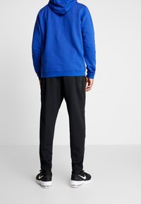 Sweat à capuche bleu avec cordon de serrage, pantalon de jogging noir avec poches latérales, et baskets noires avec semelles blanches. Tissu lisse et coupe décontractée.