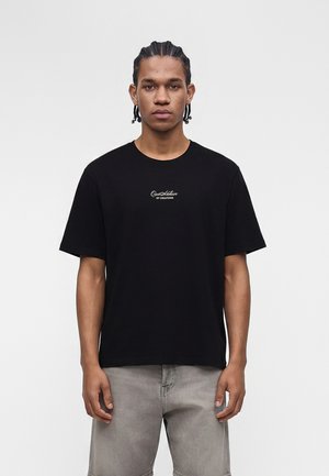 Jeune homme aux cheveux tressés portant un t-shirt noir avec un texte blanc « Que Active of Creations » et un pantalon gris délavé, debout devant un fond uni.