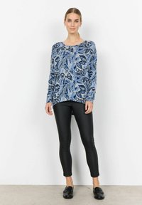 Pull bleu à motif floral avec manches longues, associé à un legging noir ajusté et des mocassins noirs, sur fond uni.