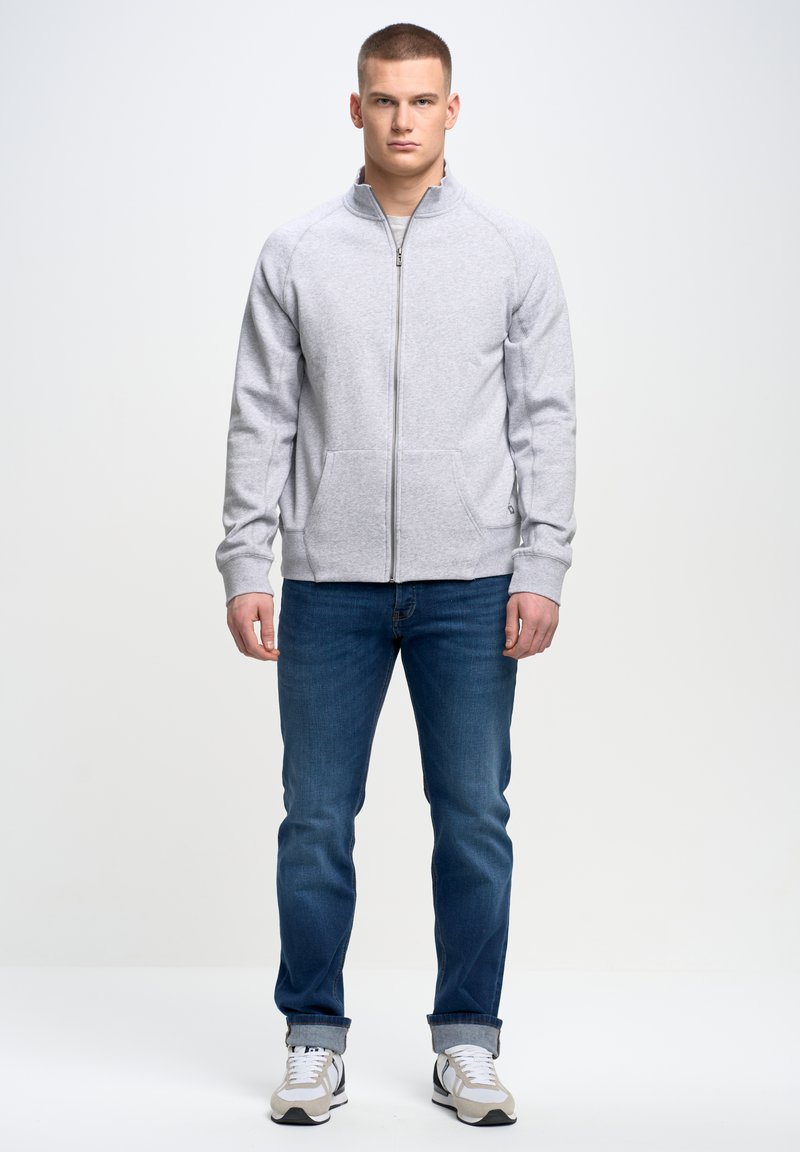 Szara bluza zapinana na zamek z wysokim kołnierzem, ściągaczami przy rękawach i kieszeniami bocznymi, w połączeniu z niebieskimi dżinsami i szarymi butami sportowymi.