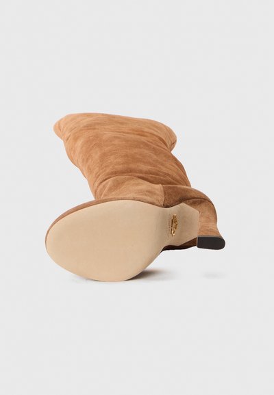 Bota de ante marrón hasta la rodilla con punta redonda y tacón afilado. La suela es de color beige con un diseño minimalista y un detalle de marca sutil.