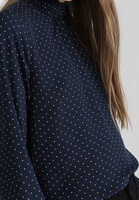 Navyblus med vita prickar, med en smockad hög halsringning och långa ärmar, tillverkad av ett mjukt, lätt material.