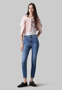 Camiseta blanca con logo, suéter de punto rosa claro colgado sobre los hombros, jeans skinny azules y zapatos negros sin cordones.