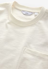 Kurzärmeliges T-Shirt in Cremefarbe mit rundem, geripptem Ausschnitt und kleinem Fronttasche. Hergestellt aus weichem, strukturiertem Stoff. Entspannter Schnitt.