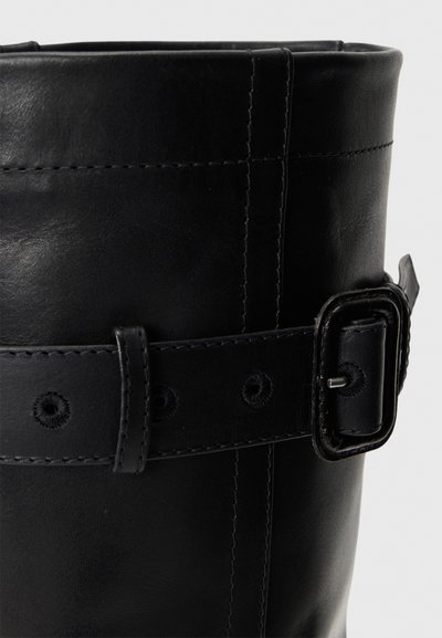 Close-up of black leather boot shaft with stitched strap and rectangular buckle fastening on a white background.