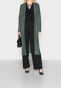 Green wool coat with straight silhouette, black blouse with tie detail, wide black pants, and green patent leather heels, complemented by a black handbag.