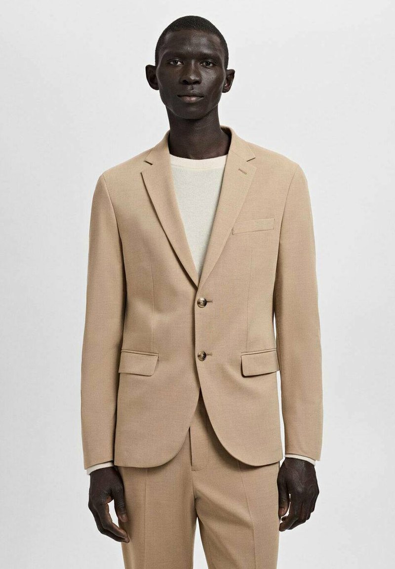 Beige suit jacket made of textured fabric, featuring a notch lapel, two buttons, and two front pockets. Worn over a light-colored top.