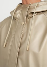 Μπεζ αδιάβροχο μπουφάν με απαλό υφή, που διαθέτει κουκούλα, κορδόνια και χρυσούς μεταλλικούς λεπτομέρειες. Ομαλή επιφάνεια και μίνιμαλ σχεδιαστικά στοιχεία.