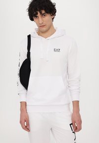 Hvid hættetrøje med EA7-logo på brystet, kængurulomme og sorte detaljer på ærmerne. Sættes sammen med hvide sweatpants.