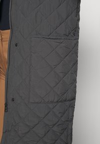 Giacca imbottita grigio scuro con un design strutturato, cuciture diagonali, chiusura con zip e una tasca visibile sul lato sinistro.