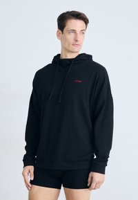 Svart hettegenser med teksturert stoff, med en rød logo på brystet. Båret med svarte tettsittende shorts. Nøytral bakgrunn.