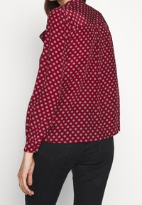 Mujer con una blusa roja de manga larga con patrones geométricos en blanco y negro, combinada con pantalones negros, vista desde atrás a la altura de la cintura.