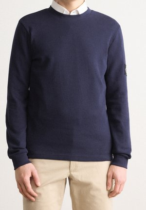 Marineblauer Langarmpullover aus strukturiertem Strickstoff. Mit einem Rundhalsausschnitt und einem Logopatch am linken Ärmel. Kombiniert mit beigen Hosen.