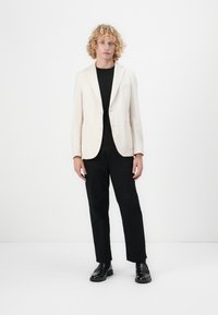 Světle béžový blazer s klopami, jednobuttunovým zapínáním, dvěma předními kapsami a černou košilí. V kombinaci s černými kalhotami a botami.