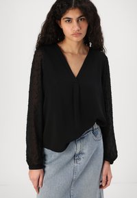 Svart bluse med v-hals og gjennomsiktige, utsmykkede lange ermer. Stoffet er glatt, og passformen er avslappet, kombinert med et denimskjørt.