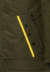 Olivgrüne Jacke mit gelben Streifen, mehreren schwarzen Knöpfen, einer Reißverschlusstasche und einer strukturierten Oberfläche.