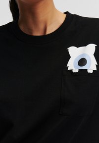 Camiseta de algodón negra con un personaje de dibujo animado blanco y de ojos azules asomándose de un bolsillo en el pecho. Diseño básico de cuello redondo.