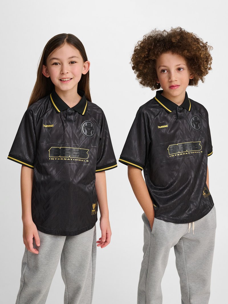 Zwei Kinder tragen schwarze Sporttrikots mit gelbem Saum und graue Jogginghosen und stehen vor einem weißen Hintergrund.