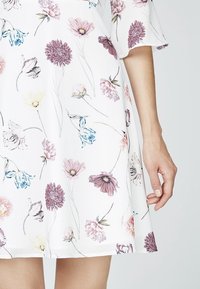 Blommig klänning med vit bakgrund; har lila, rosa och blå blommor. Korta ärmar, A-linjeformad siluett, lätt tyg.