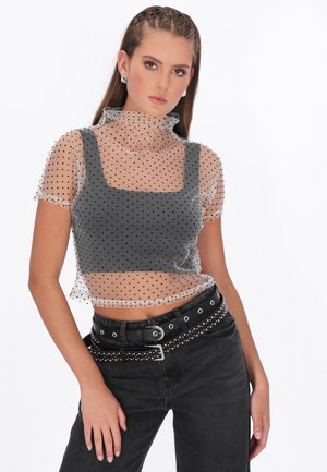 Jonge vrouw met vlechten, gekleed in een doorschijnend topje met polkadots over een zwart crop topje en zwarte spijkerbroek met studs en dubbele riemen.
