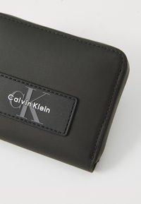 Zwarte portemonnee met een gladde textuur, voorzien van een rechthoekig logo patch met "Calvin Klein" in witte tekst. Strak ontwerp met gestikte randen.