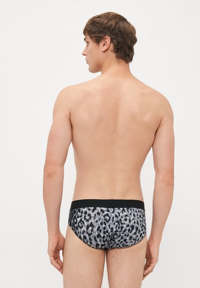 Jeune homme aux cheveux bruns courts, de dos, torse nu, portant un slip léopard gris et noir avec une ceinture noire unie.