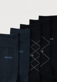 Cinq paires de chaussettes dans des tons foncés, avec une texture côtelée, un détail de logo, et une paire avec un motif losange gris. Différentes nuances de marine et de noir.