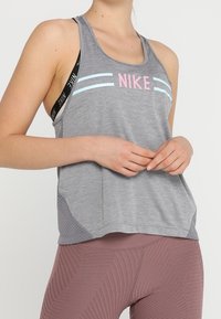 Canotta sportiva grigia con strisce azzurre e rosa, realizzata in un tessuto traspirante con pannelli laterali in rete, indossata sopra leggings a fantasia.