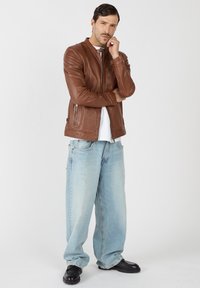 Braune Lederjacke mit Stehkragen, Frontreißverschlüssen und lockerer Passform. Kombiniert mit hellblauen, oversized Jeans und schwarzen Schuhen.