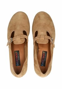 Zapatos de ante en color marrón claro con puntera redondeada, detalles recortados y una correa ajustable con hebilla. La plantilla presenta una etiqueta de marca colorida.