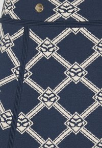 Tejido de color azul marino con un diseño de patrón de diamantes en crema. Presenta un botón de acento metálico y una superficie texturizada que destaca formas gráficas.