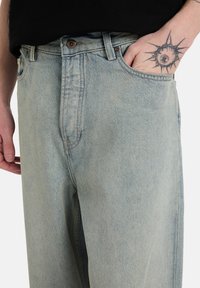 Τζιν παντελόνια από ανοιχτό μπλε ντενίμ με κλασικό σχεδιασμό πέντε τσεπών, με χάλκινα μεταλλικά στοιχεία και ελαφρώς ξεθωριασμένη υφή. Χέρι εν μέρει μέσα στην τσέπη.